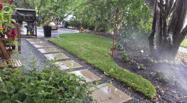 A wet stone path leads through a green garden with a grill, torches, and flowers. A sprinkler waters the grass, and trees provide shade.