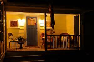 A warmly lit front porch at night features a blue door, two chairs, a small table with a plant, and a railing in the foreground. The house exterior is yellow.