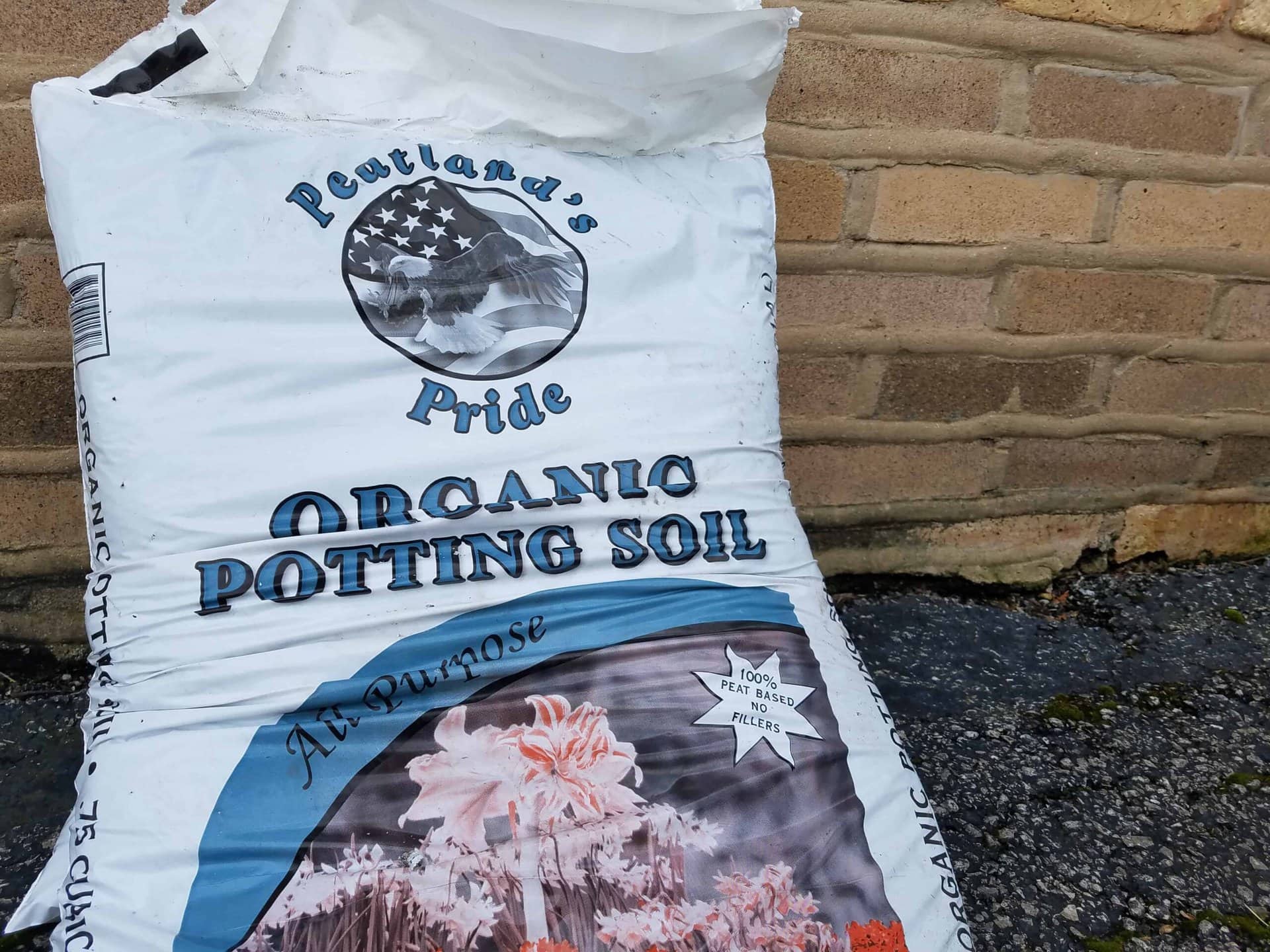 A white bag labeled Peatlands Pride Organic Potting Soil rests against a brick wall on asphalt. The bag features an American flag and plant graphics.
