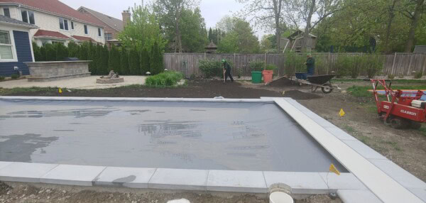 A backyard under construction with a covered pool in the foreground, workers near a wheelbarrow, a patio area, green plants, and a wooden fence in the background.