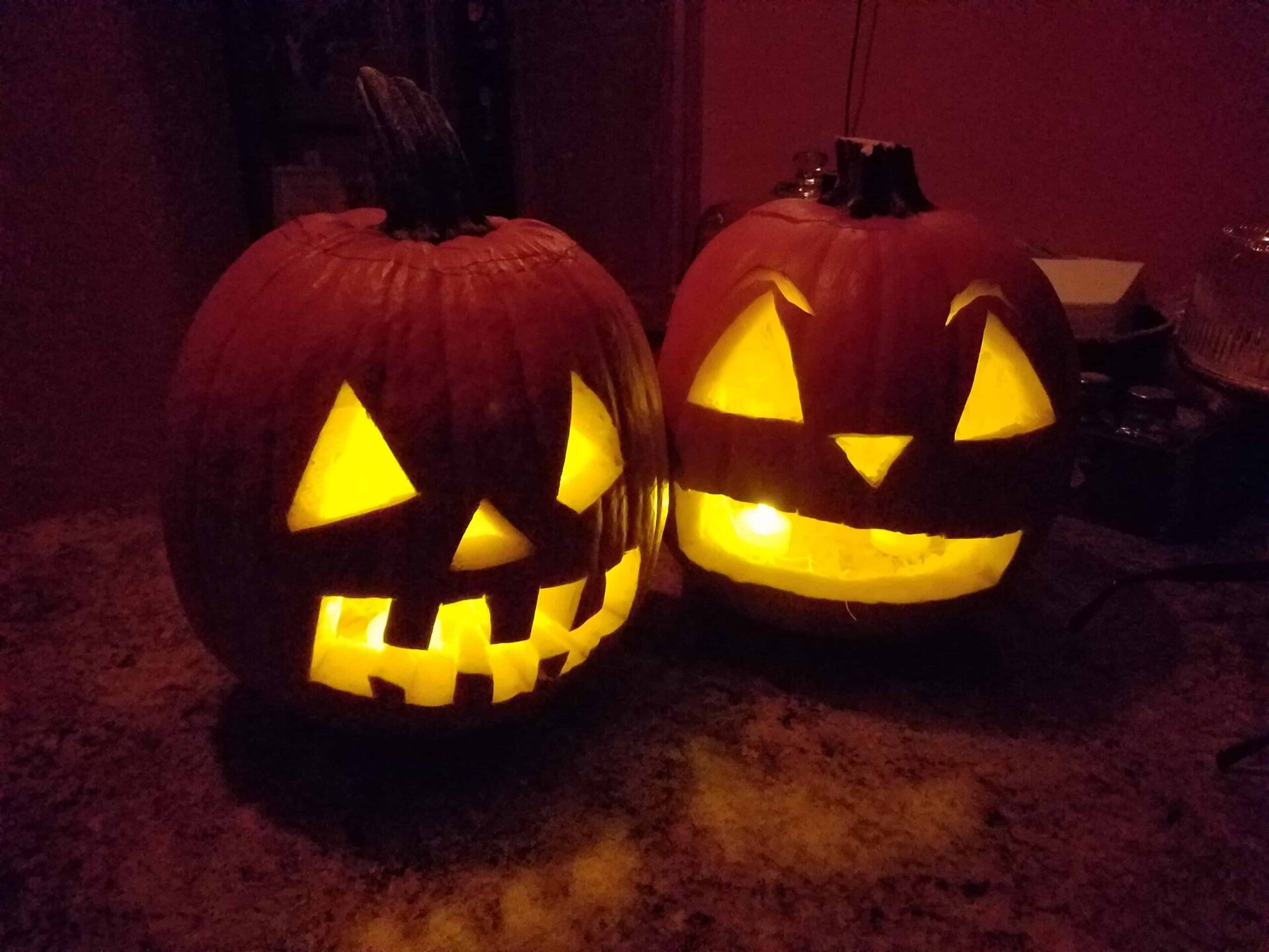 Two carved jack-o-lantern pumpkins with glowing faces sit on a dark surface. One pumpkin has a toothy grin and triangular eyes; the other has wide eyes and a big smile. Both are lit from inside, creating a warm glow.