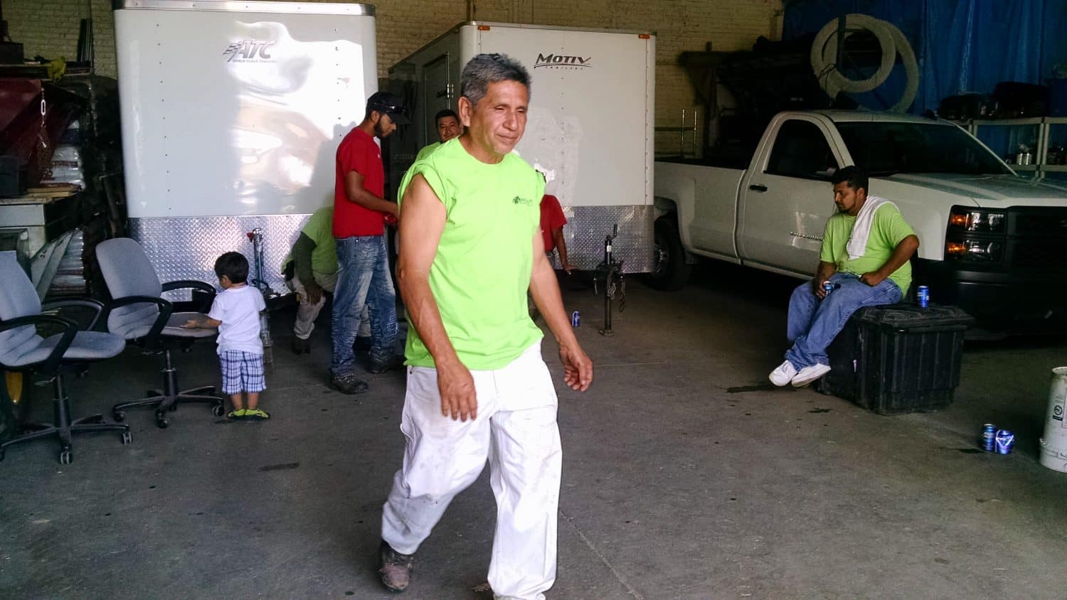 A man in a neon green shirt and white pants walks indoors, smiling. Behind him, several people sit or stand near trailers and chairs, and a child stands by an adult. A white truck is parked on the right.