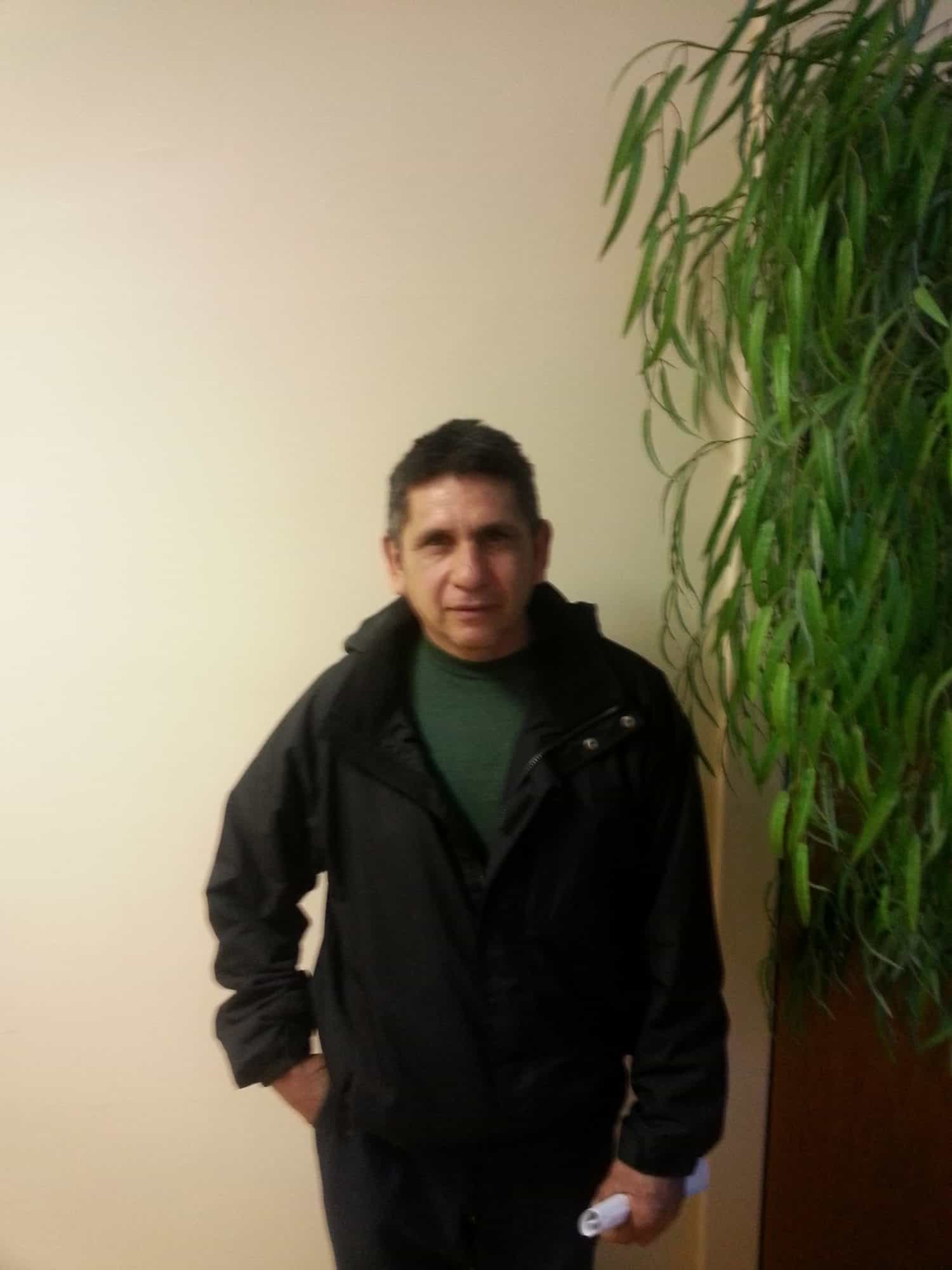 A man wearing a black jacket stands indoors next to a tall green plant, with a beige wall in the background. He has short dark hair and is holding something white in his right hand.