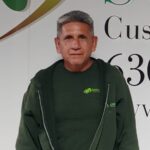 A man with gray hair in a green SWDI shirt and hoodie stands in front of a wall with part of a company logo and phone number visible in the background.