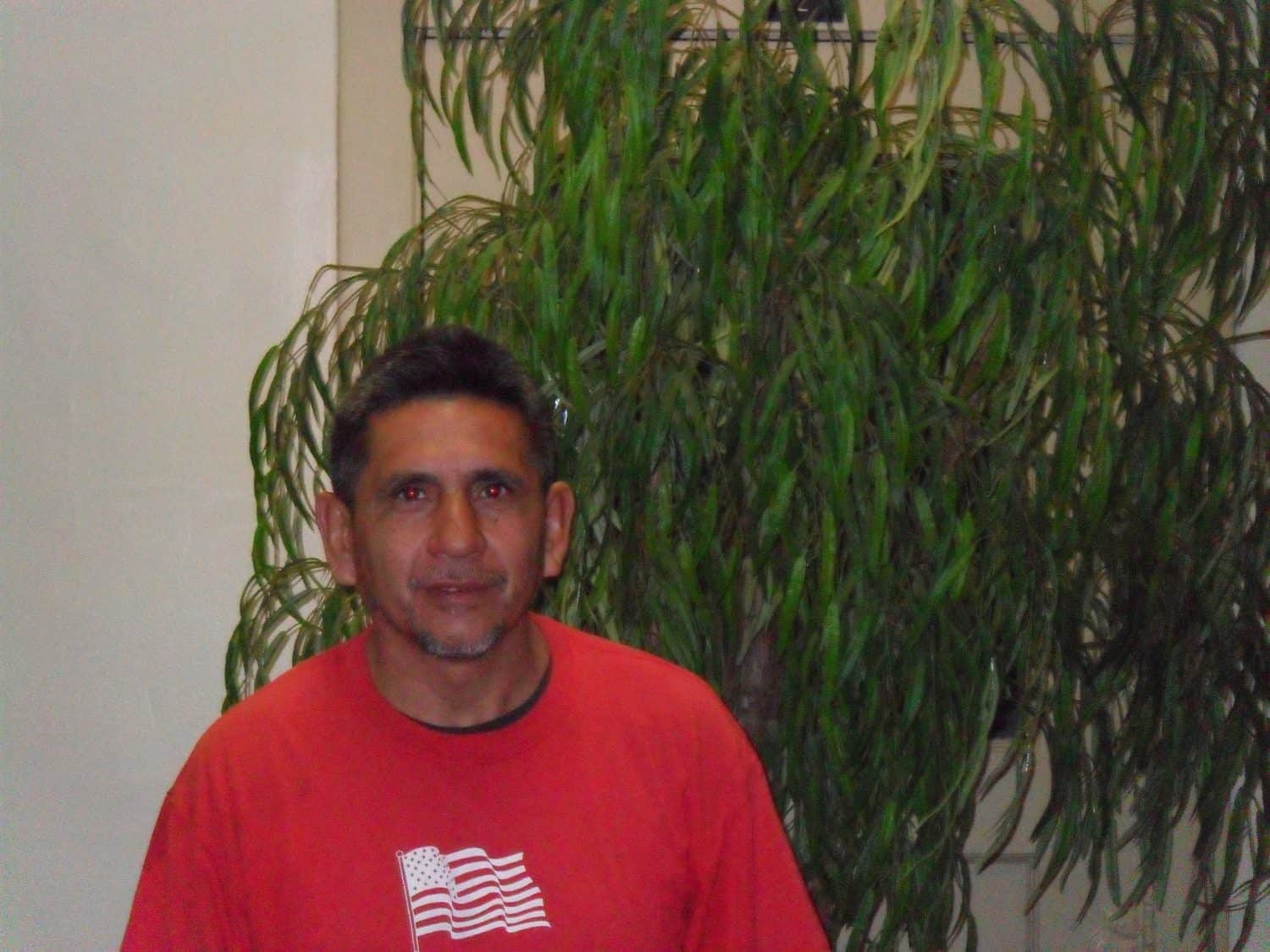Rogelio D. A man in a red shirt with an American flag design stands in front of a tall green leafy plant indoors.