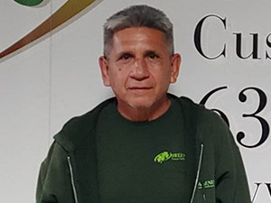 A man with short gray hair wearing a green hoodie and green t-shirt stands in front of a white wall with partially visible text and numbers.