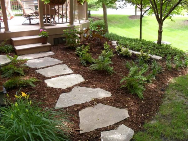 Stepping stones form a path through a mulched garden with ferns and shrubs, leading to a porch with wooden steps and potted plants. Trees and lawn are in the background.