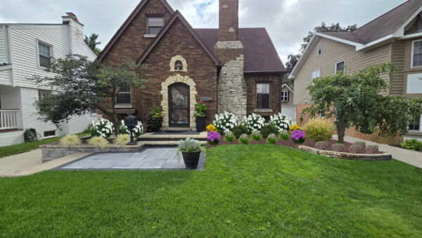 A brick house with a stone chimney, surrounded by colorful flowers, shrubs, and green lawn. Theres a paved patio area with a potted plant by the front entrance. Neighboring houses are visible on both sides.
