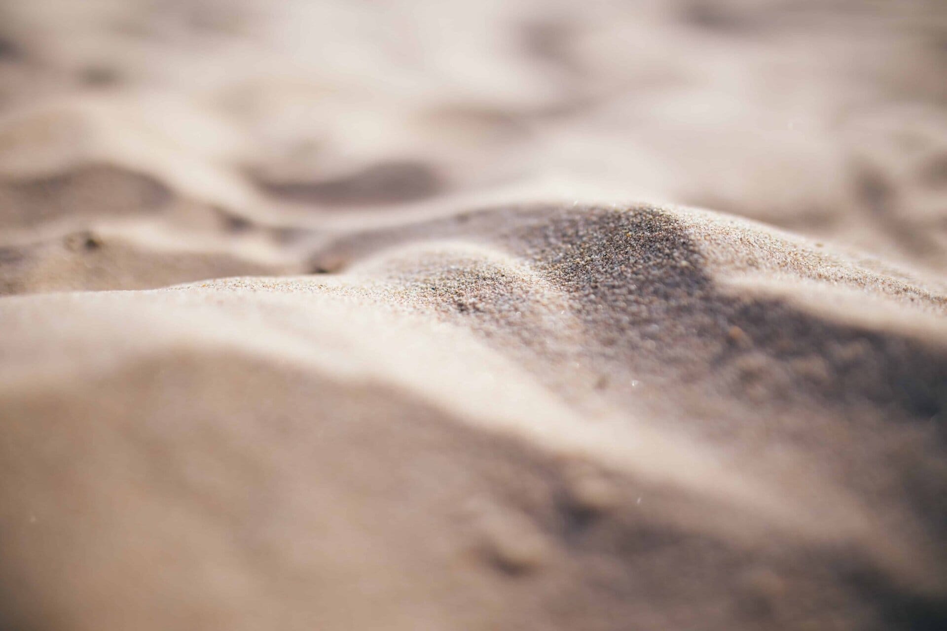Close-up of soft, sunlit sand with gentle, uneven mounds and a blurred background, capturing the texture and tiny grains in the foreground.