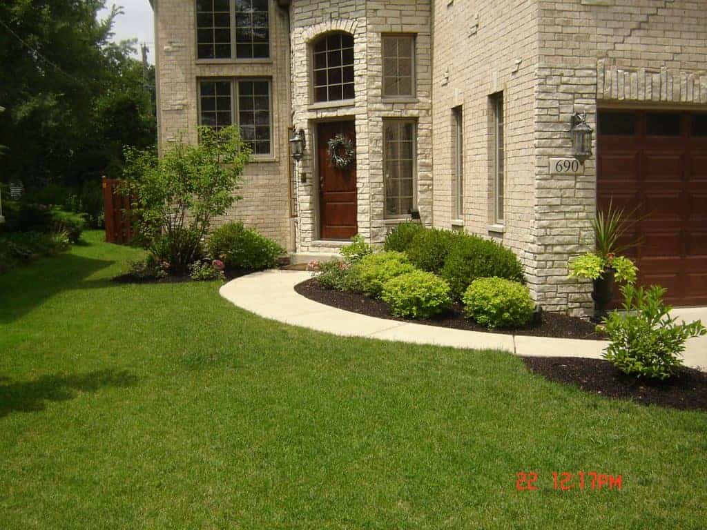 A well-maintained front yard with green grass, neatly trimmed bushes, and a curved sidewalk leading to the wooden front door of a light brick house.