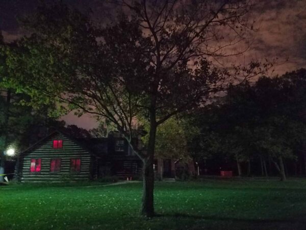 A log cabin with red-lit windows sits behind a tree at night, surrounded by a grassy yard and dense, shadowy trees under a cloudy, dimly lit sky.