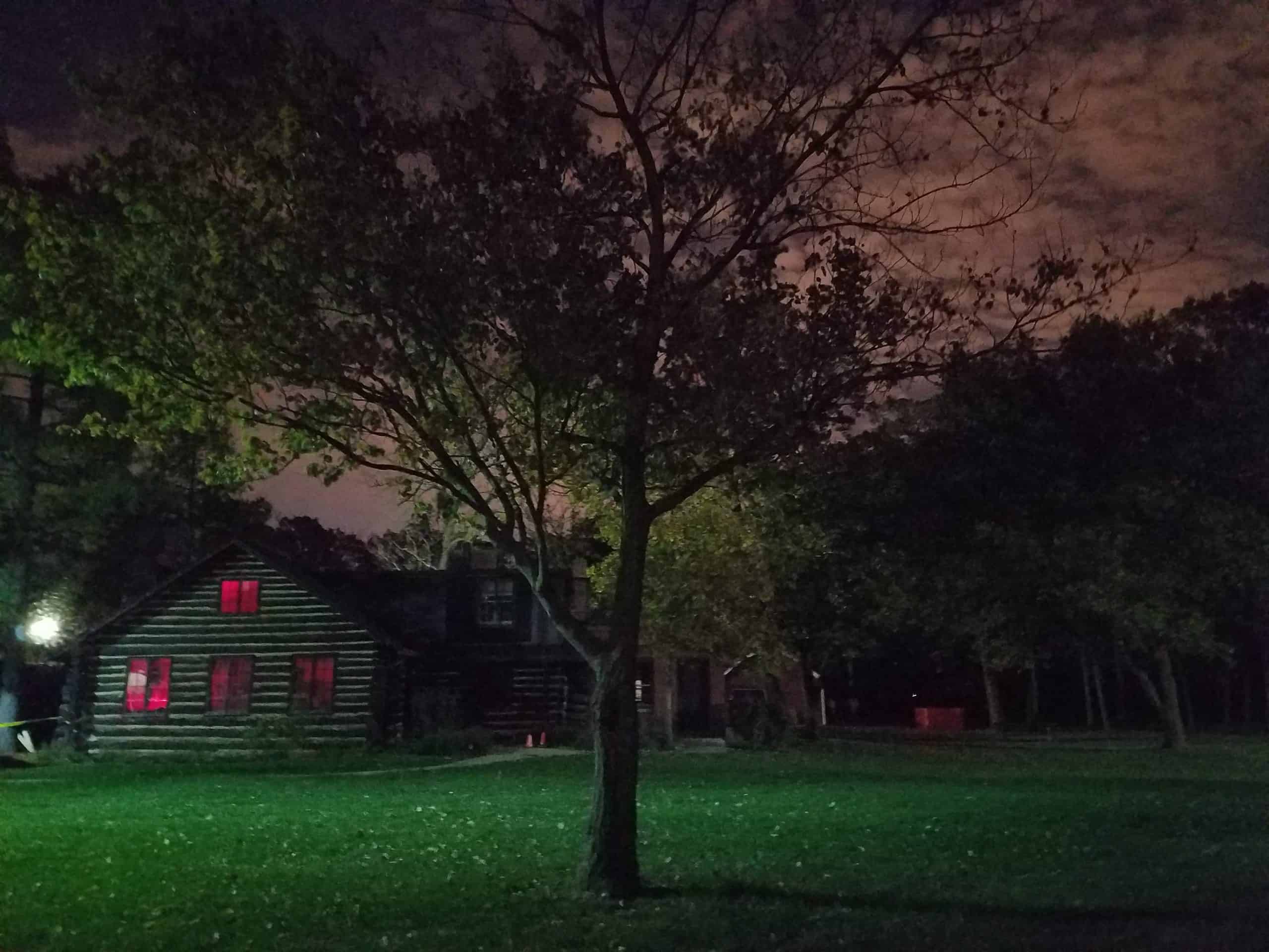 A log cabin with red-lit windows sits behind a tree at night, surrounded by a grassy yard and dense, shadowy trees under a cloudy, dimly lit sky.