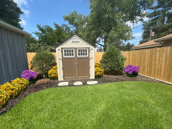 A small tan and brown shed is centered in a fenced backyard, surrounded by green shrubs and colorful flowers on fresh mulch with stepping stones leading to the door. Lush grass and a sunny blue sky complete the scene.