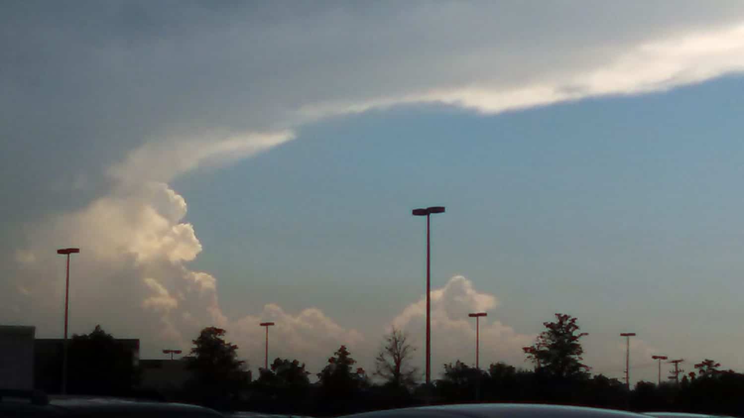 Large, dramatic clouds roll across a blue sky above a parking lot with silhouetted cars, trees, and tall light poles.