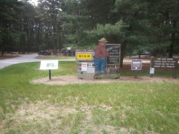 A forest entrance sign displays Todays Fire Danger: HIGH with Smokey Bear, warning visitors to prevent forest fires. Several informational signs are visible. Cars and trees are in the background.
