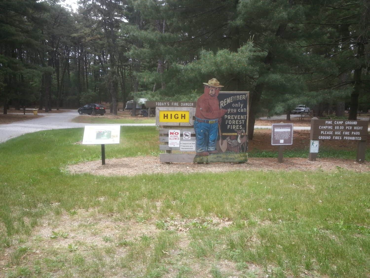 A Smokey Bear fire danger sign shows todays fire danger level as HIGH at a forest campground. Several signs with safety information are nearby, with parked cars and trees in the background.