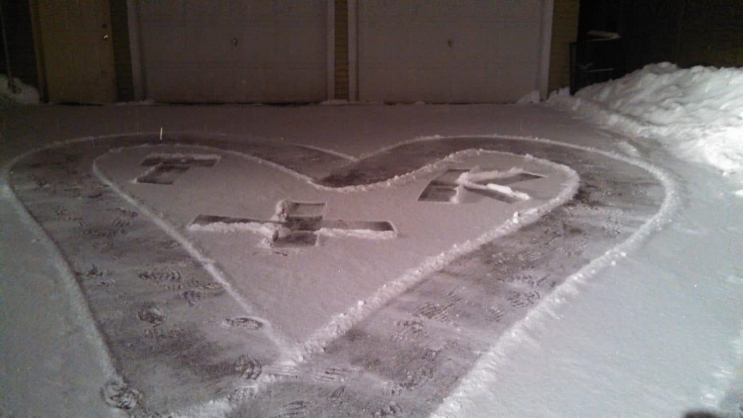 A large heart shape is etched in fresh snow on a driveway, with the letters T + R inside. Snow piles line the edges, and garage doors are visible in the background.