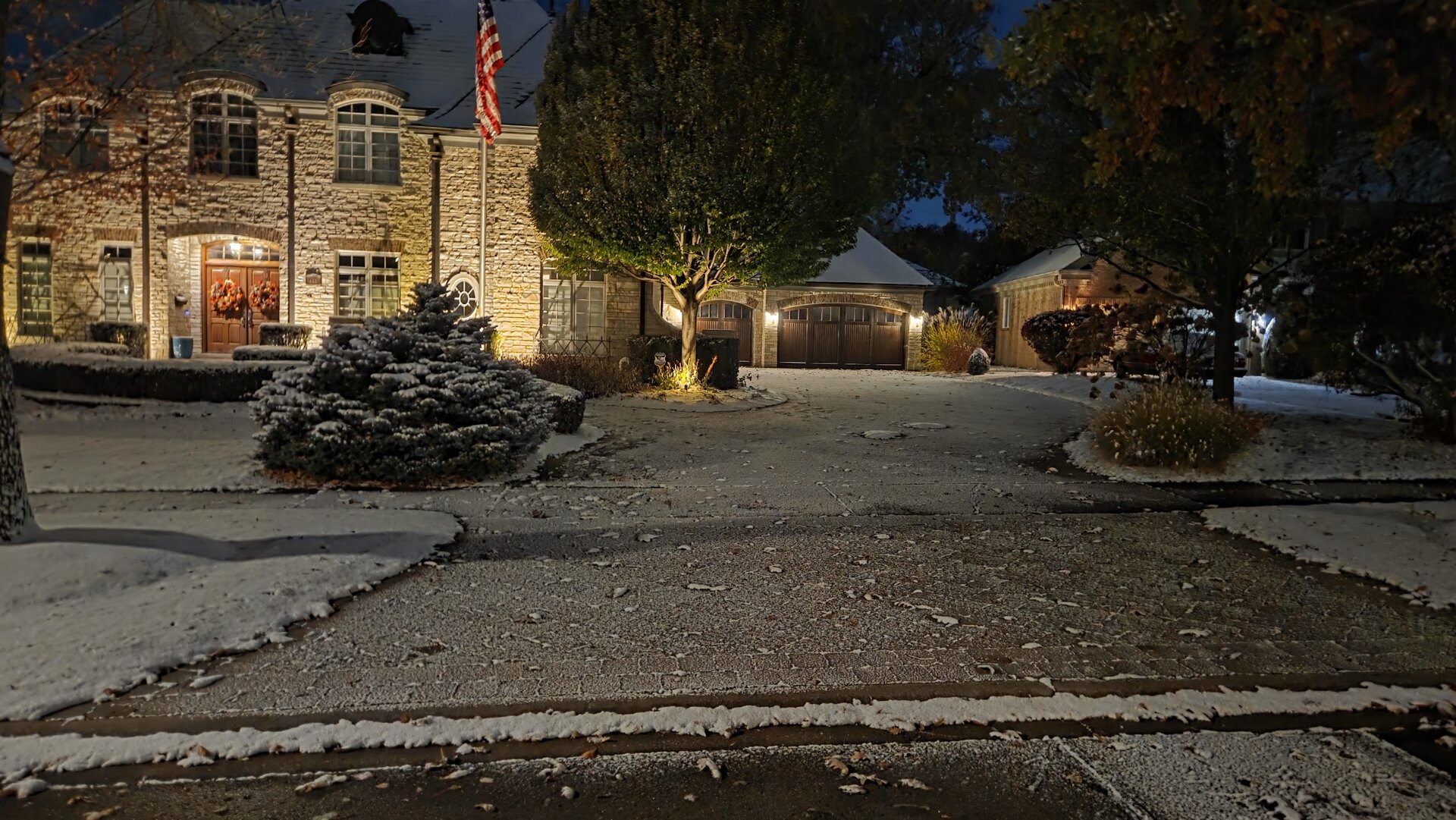 A large house with stone walls, outdoor lights, and an American flag stands by a driveway lightly dusted with snow at night. Trees and bushes surround the home.