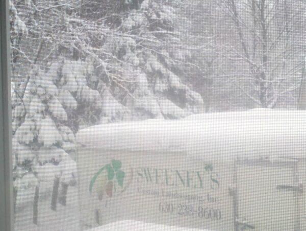 A snow-covered trailer with Sweeneys Custom Landscaping Inc. and a phone number is parked in front of snow-laden trees during heavy snowfall.