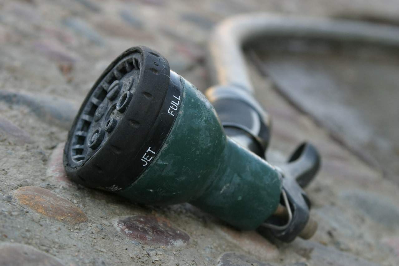 A green garden hose nozzle lies on a textured, concrete surface with a hose attached, slightly out of focus in the background.