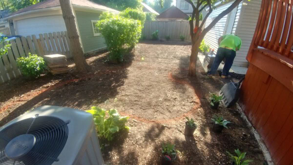 A person prepares a backyard garden, digging near a house. Potted plants, a red deck, air conditioning unit, and trees are visible. Sunlight filters through the trees and fence.