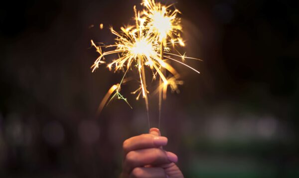A hand holding two lit sparklers with bright sparks against a dark, blurred background.