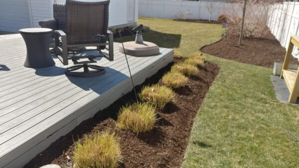 A backyard with a wooden deck, swivel chair, and side table. Trimmed bushes line the mulched garden bed next to the deck. A lawn and a white fence are in the background.