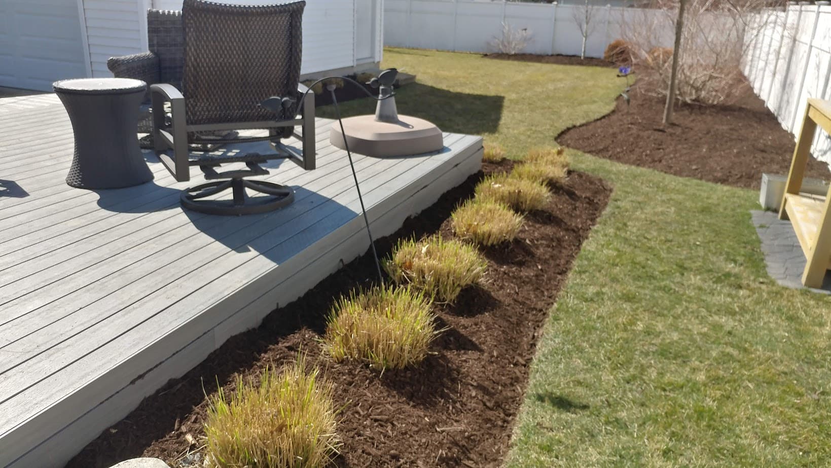 A backyard with a wooden deck, swivel chair, and side table. Trimmed bushes line the mulched garden bed next to the deck. A lawn and a white fence are in the background.