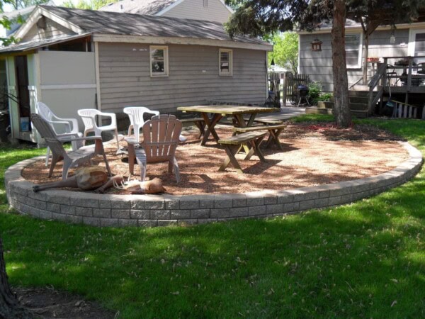 A backyard patio with picnic tables, Adirondack chairs, and white plastic chairs on gravel, bordered by a low stone wall, with grass and two houses in the background.