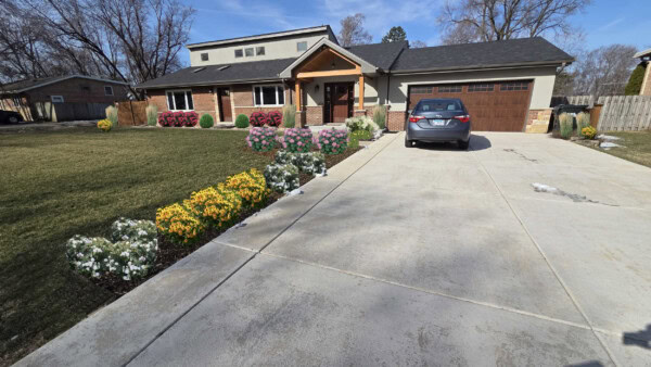 A modern house with a front porch, driveway, and a car parked. The yard features green grass, blooming yellow, white, and pink flowers, and small bushes. Trees without leaves are visible in the background.