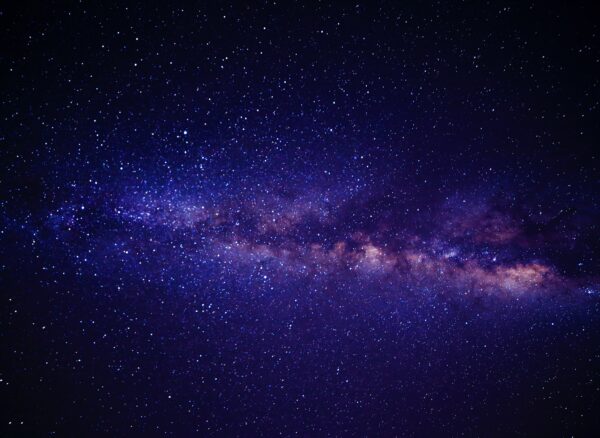 A night sky filled with countless stars and a bright, dense stretch of the Milky Way galaxy glowing across the center, set against a deep blue and purple backdrop.