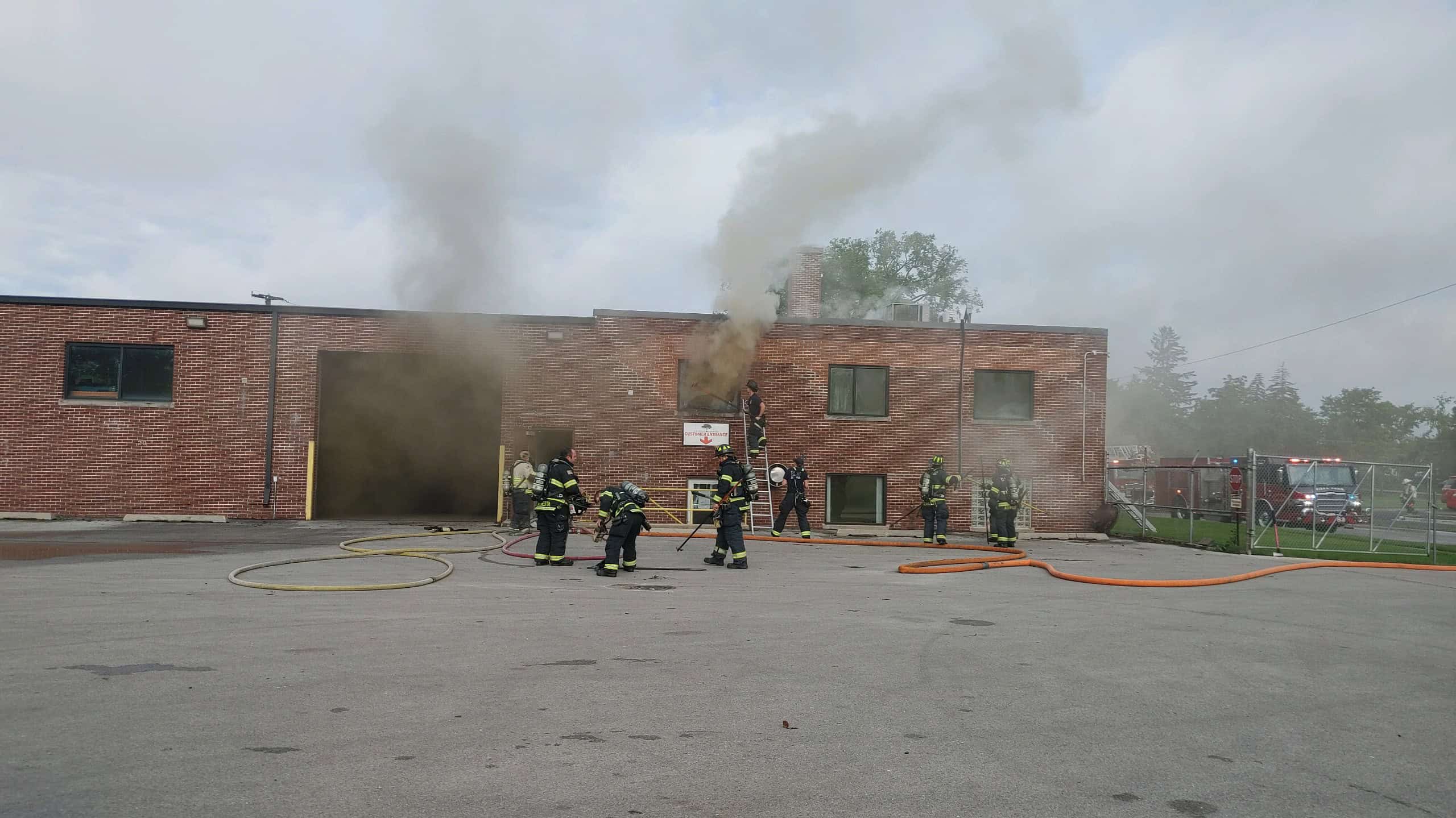 Firefighters respond to smoke pouring from the upper windows of a two-story brick building. Multiple hoses are on the ground, and a fire truck is visible to the right.