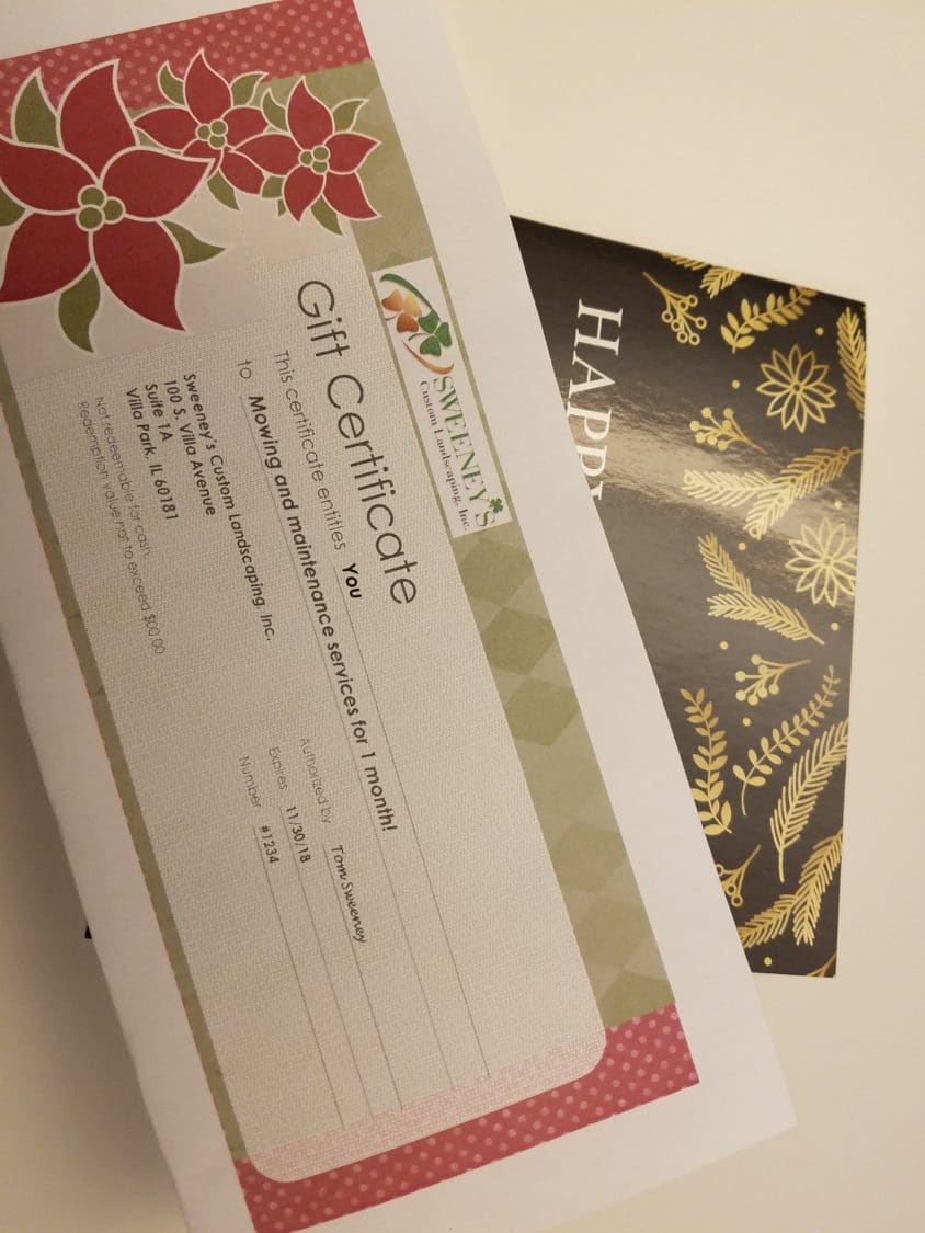 A gift certificate for one month of mowing and maintenance services is placed on top of a black card with gold floral designs and partial HAPPY text visible.