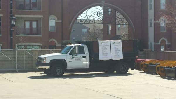 A white Habitat for Humanity truck is parked in a sunlit lot near an iron arch, with banners displayed on its side. Nearby, yellow and orange bins line the pavement. Apartment buildings are visible in the background.