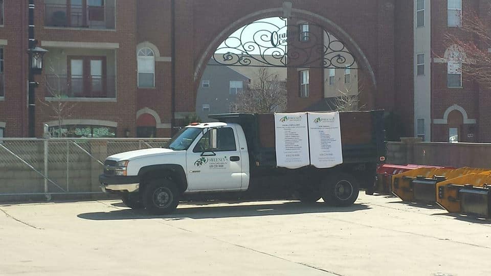 A white Habitat for Humanity truck is parked in a sunlit lot near an iron arch, with banners displayed on its side. Nearby, yellow and orange bins line the pavement. Apartment buildings are visible in the background.