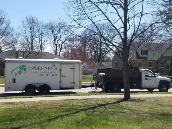 A landscaping company truck and trailer, labeled Sweeneys Custom Landscaping, Inc., are parked on a suburban street in front of brick houses and leafless trees on a sunny day.