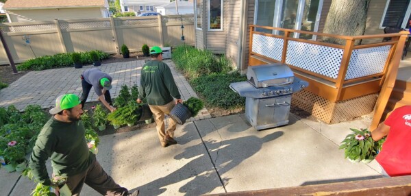Four people are gardening in a backyard. Two are carrying potted plants, one is kneeling near bushes, and another stands holding a plant. A grill and wooden deck are on the right, and sunlight casts shadows on the patio.