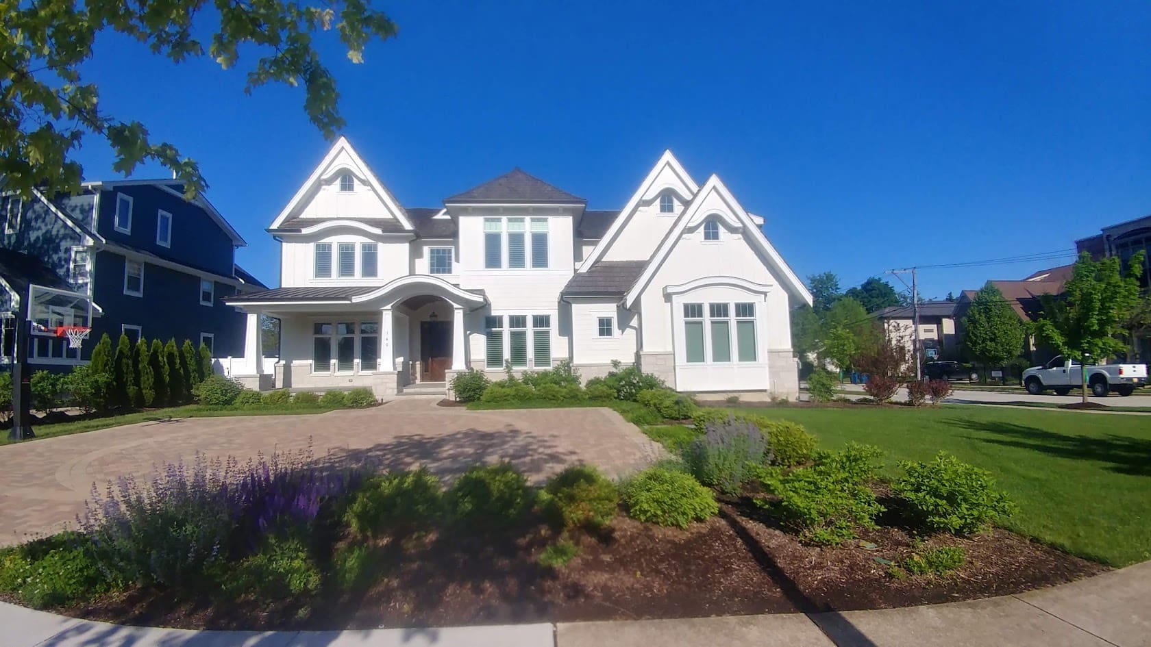 A large white two-story house with gabled roofs, a wide driveway, landscaped front yard, and green grass under a clear blue sky.