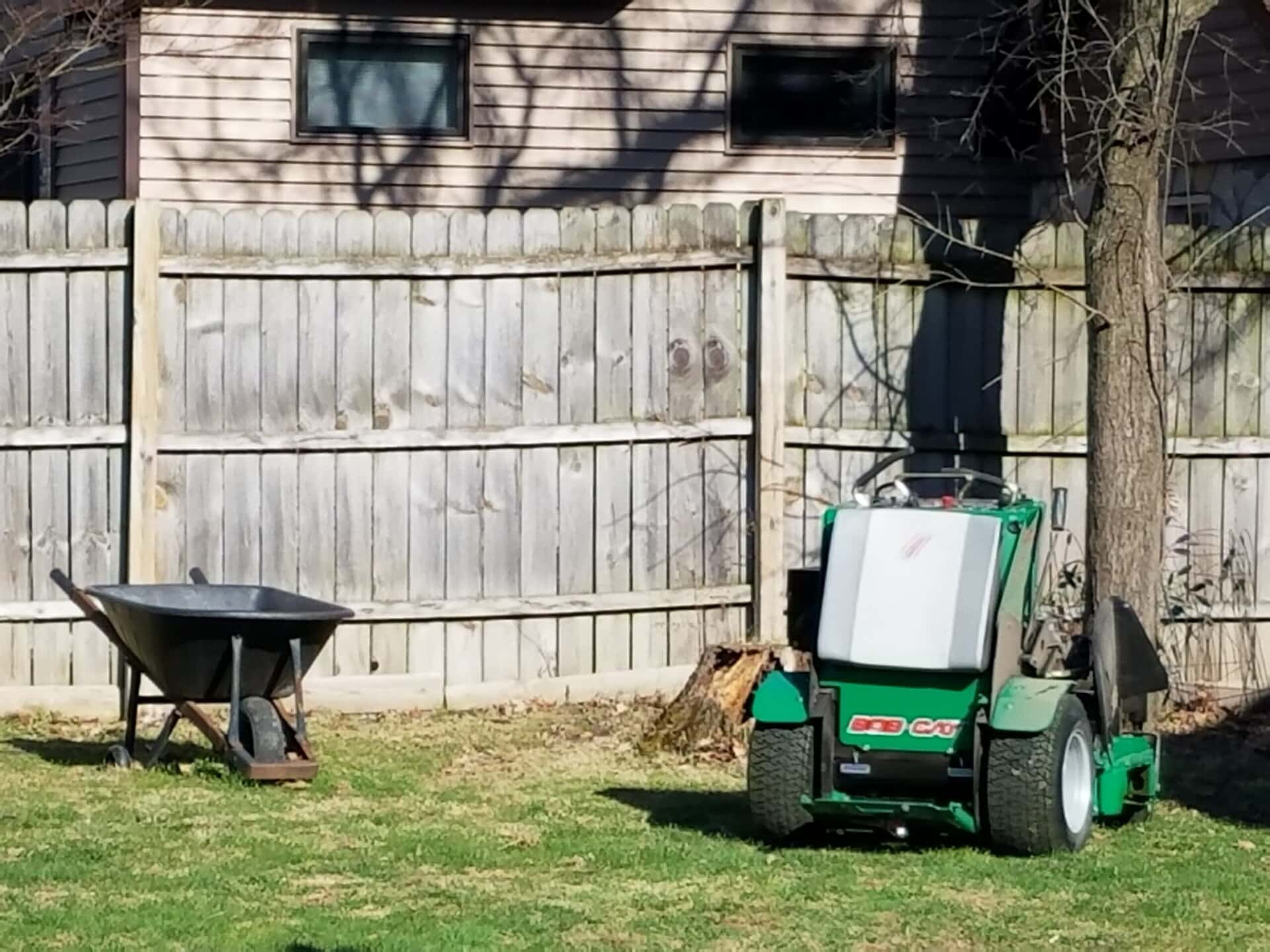 A green lawn mower and a black wheelbarrow sit on grass near a wooden fence, with a house and tree in the background.