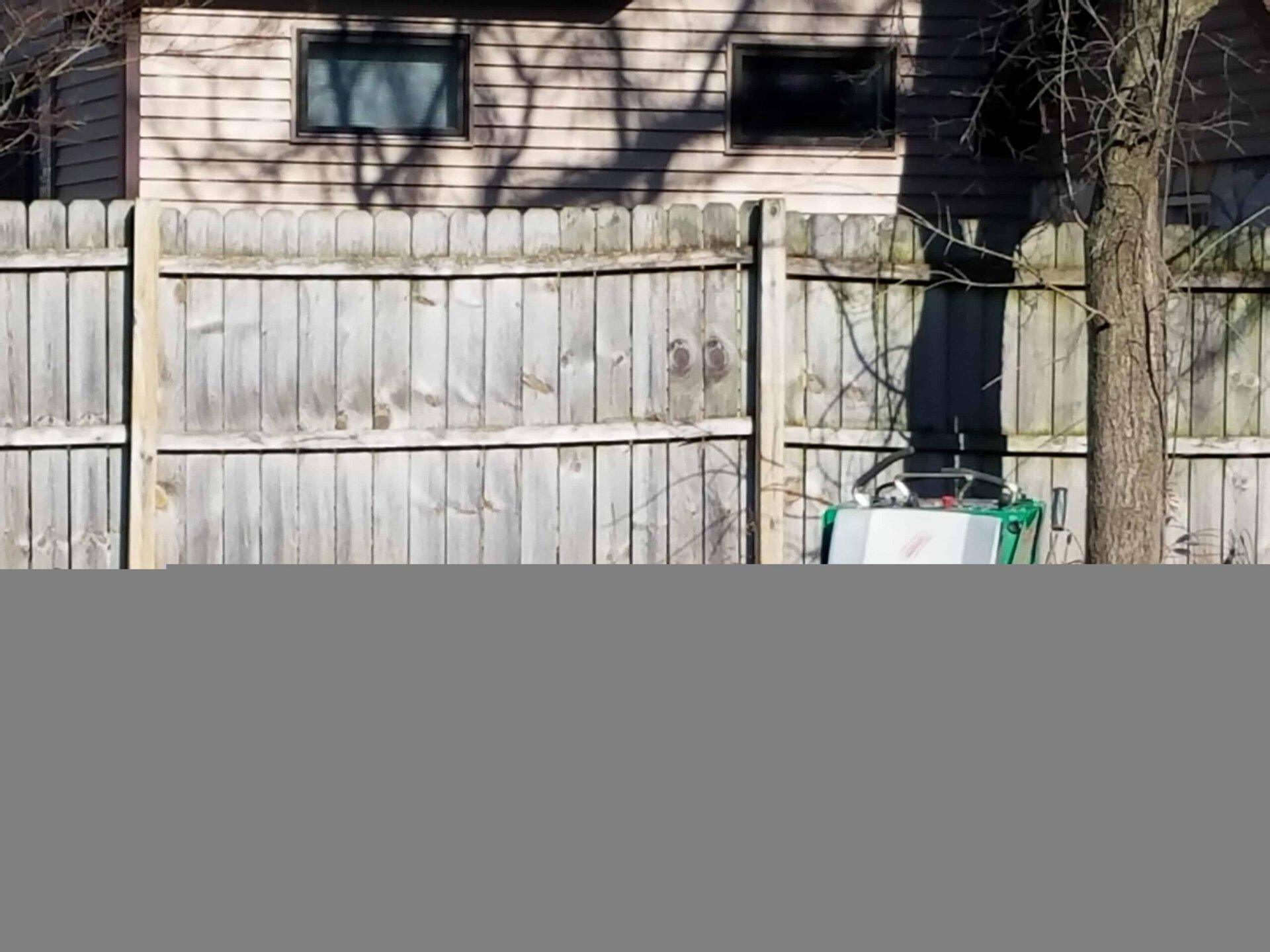 A wooden fence stands in front of a beige house with two small windows. A tree trunk and a green trash bin are visible beside the fence. Shadows from tree branches fall across the house and fence.