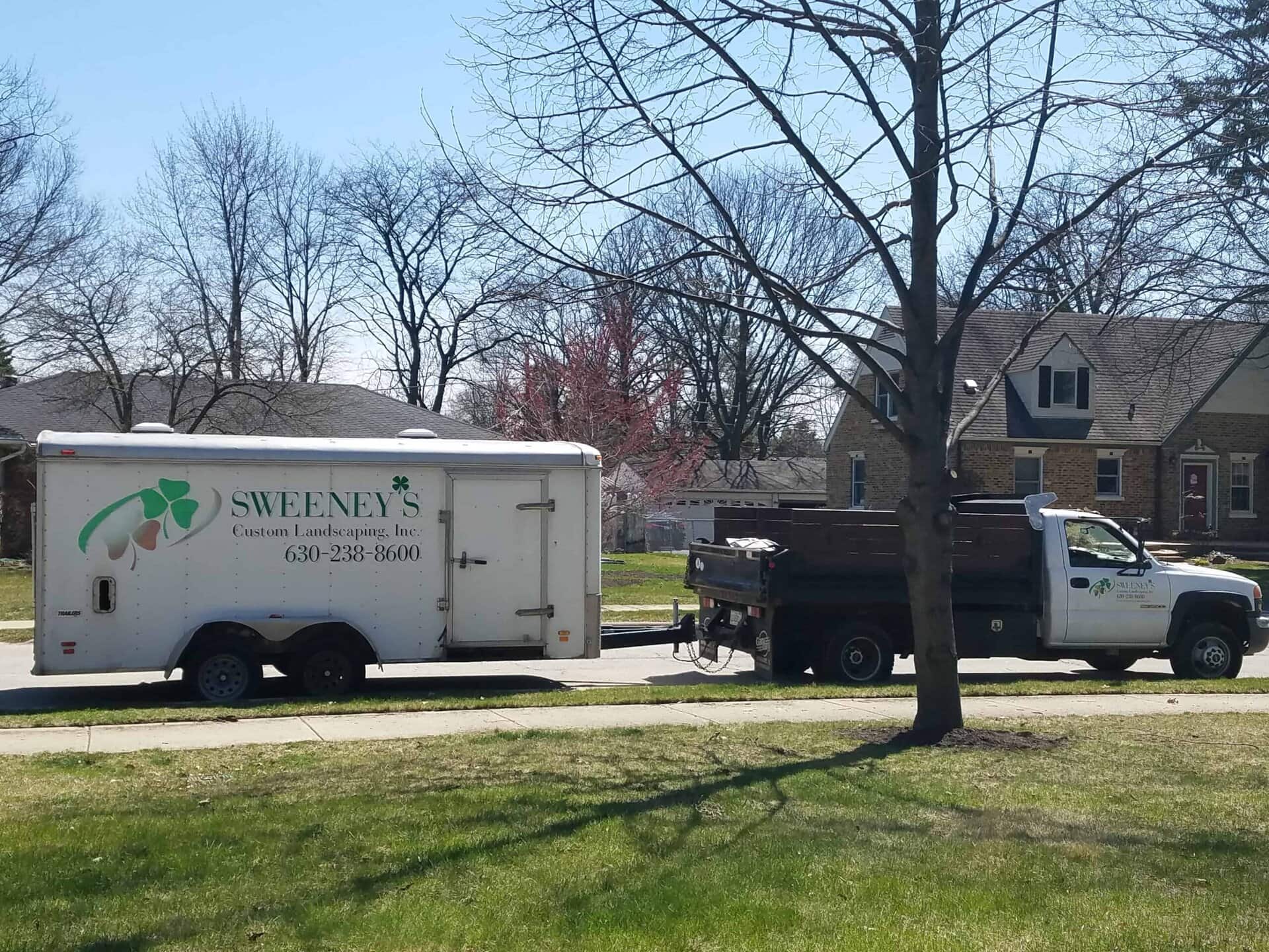 A white landscaping truck and trailer labeled Sweeneys Custom Landscaping, Inc. are parked on a suburban street near houses and leafless trees on a sunny day.