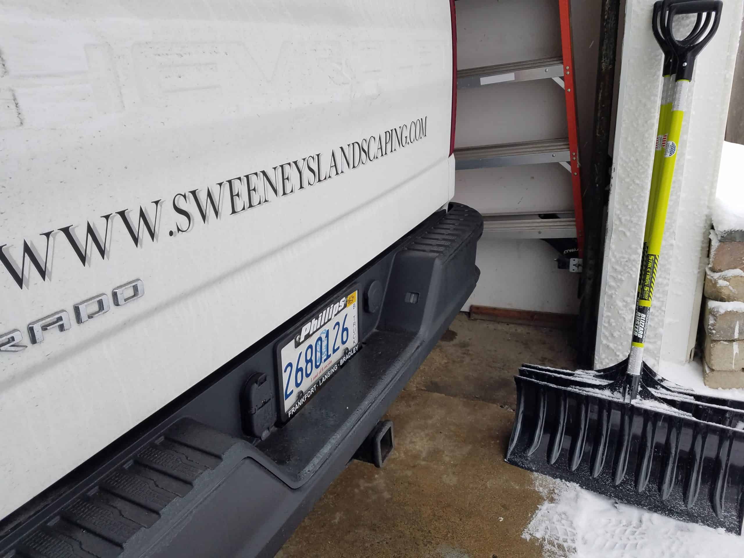 Rear of a white landscaping truck with a visible website, parked by a snow shovel, some snow on the ground, and a metal shelf in the background.