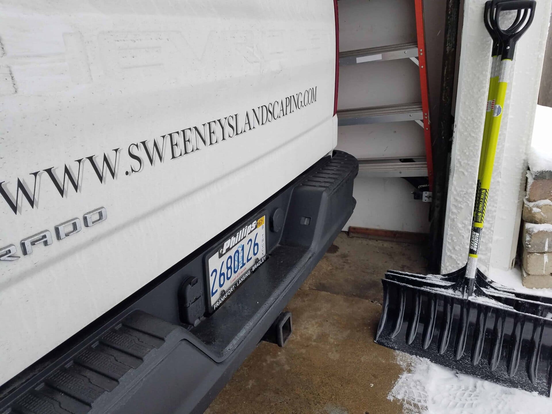 A snow shovel leans against a wall near the rear of a white truck labeled Sweeneys Landscaping, parked in a snowy driveway.