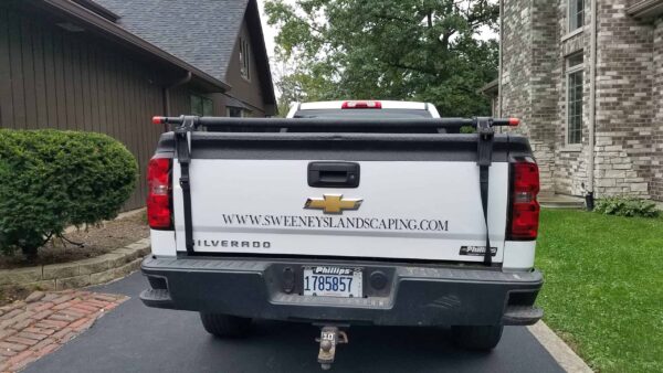 A white Chevrolet Silverado truck with the website www.sweeneylandscaping.com on the tailgate is parked in a driveway next to a brick house.