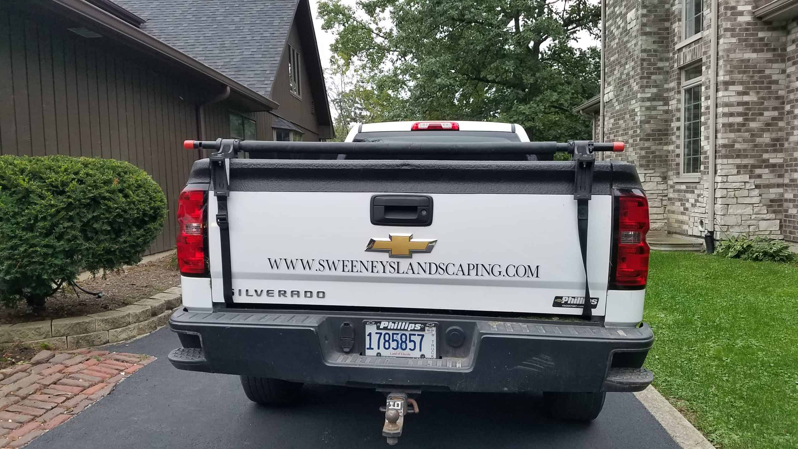 Rear view of a white Chevrolet Silverado pickup truck with www.sweenyslandscaping.com on the tailgate, parked in a driveway next to a brick house.