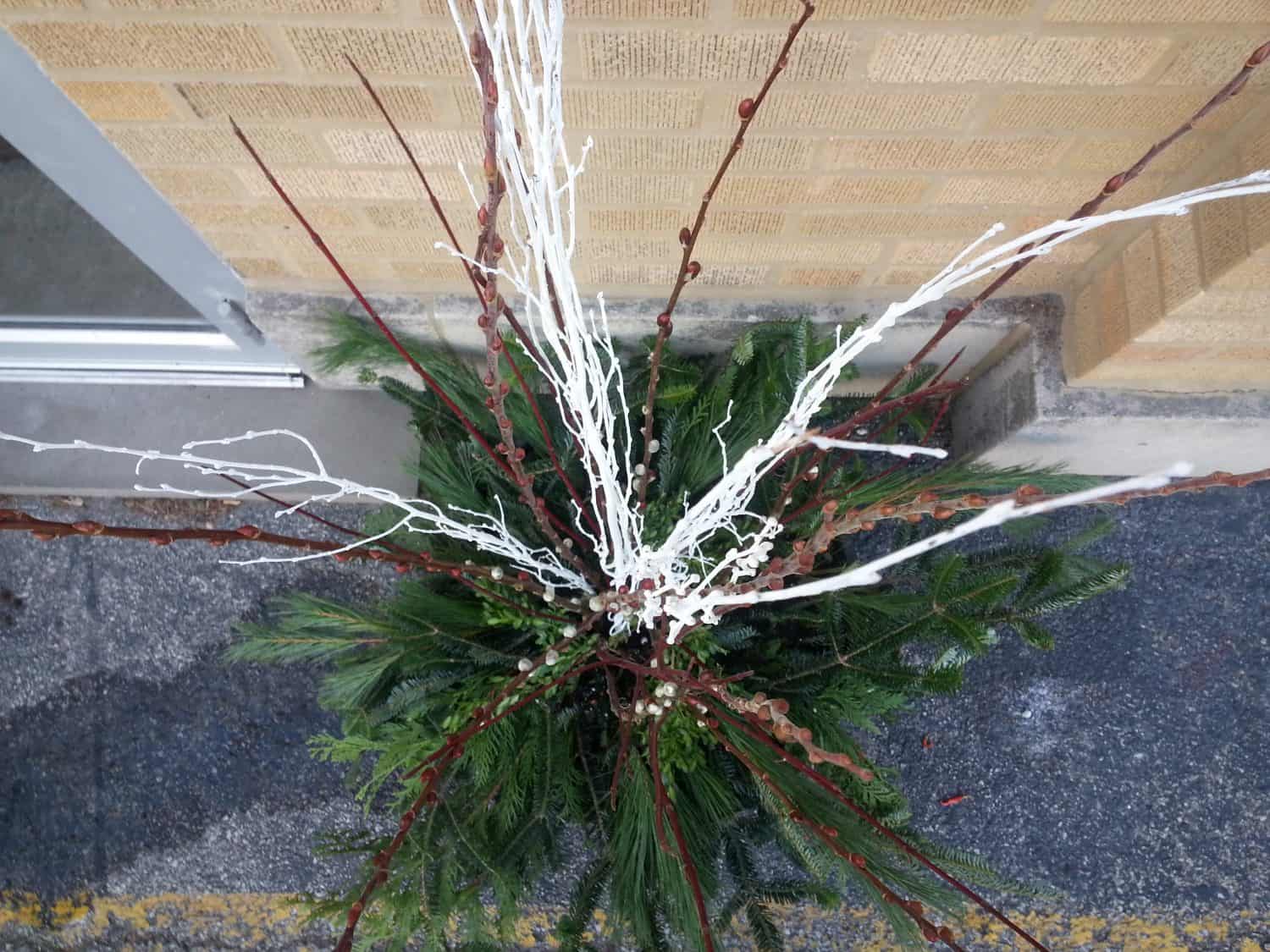 A planter with green pine branches, brown twigs, and several tall, white-painted branches is placed near a brick wall on a concrete surface.