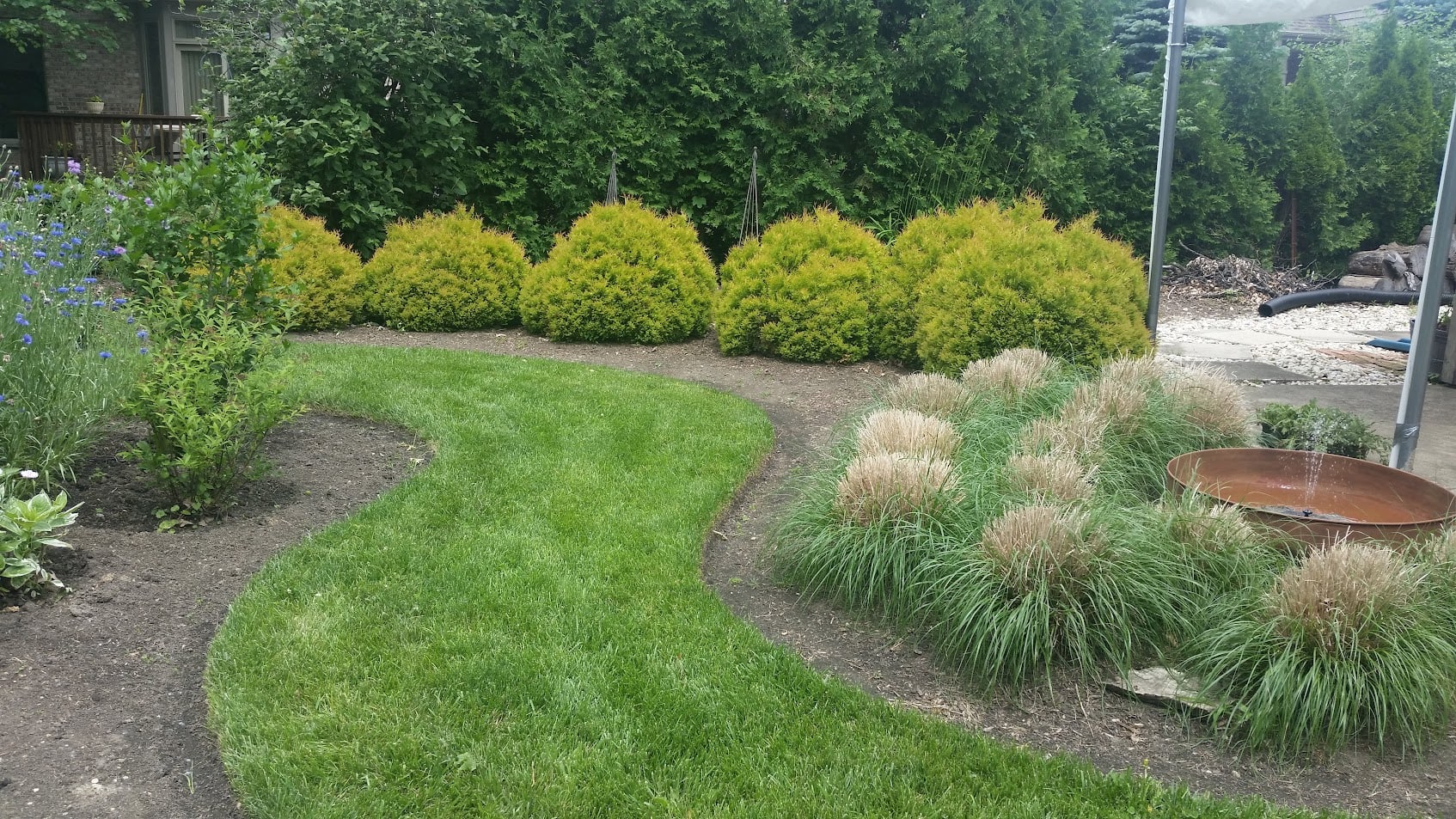 A green, curved lawn path winds between bushes and ornamental grass, with a round metal water feature on the right and dense trees and shrubs lining the background.