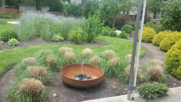 A garden with a round metal fountain in the center, surrounded by tall grasses and lush landscaping, including flower beds, shrubs, and a manicured lawn. A metal pole is visible in the foreground.