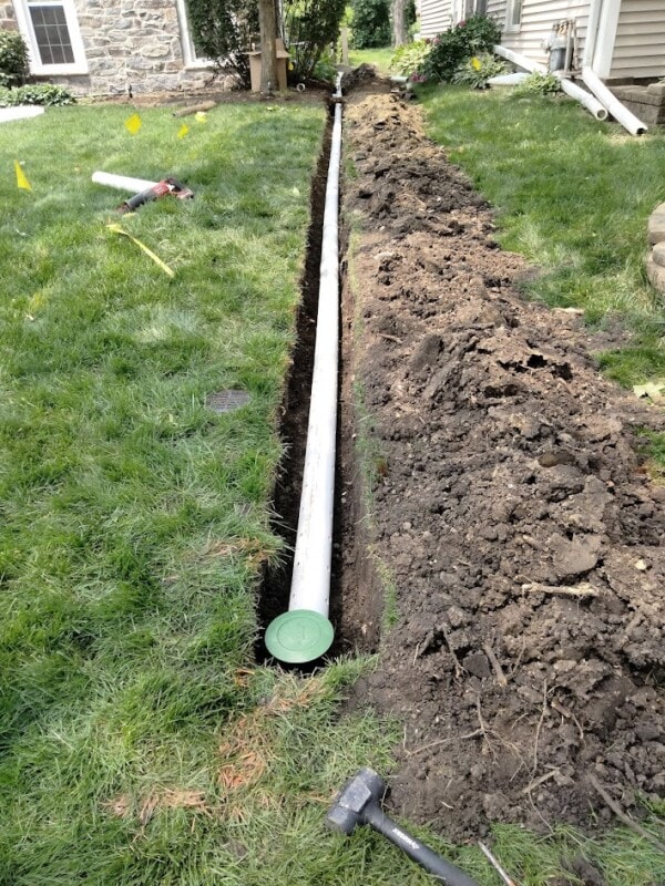 A white drainage pipe is laid in a narrow trench through a grassy yard, with soil piled alongside. A green cap is visible on one end, near a house and stone wall. Tools and yellow flags are scattered nearby.