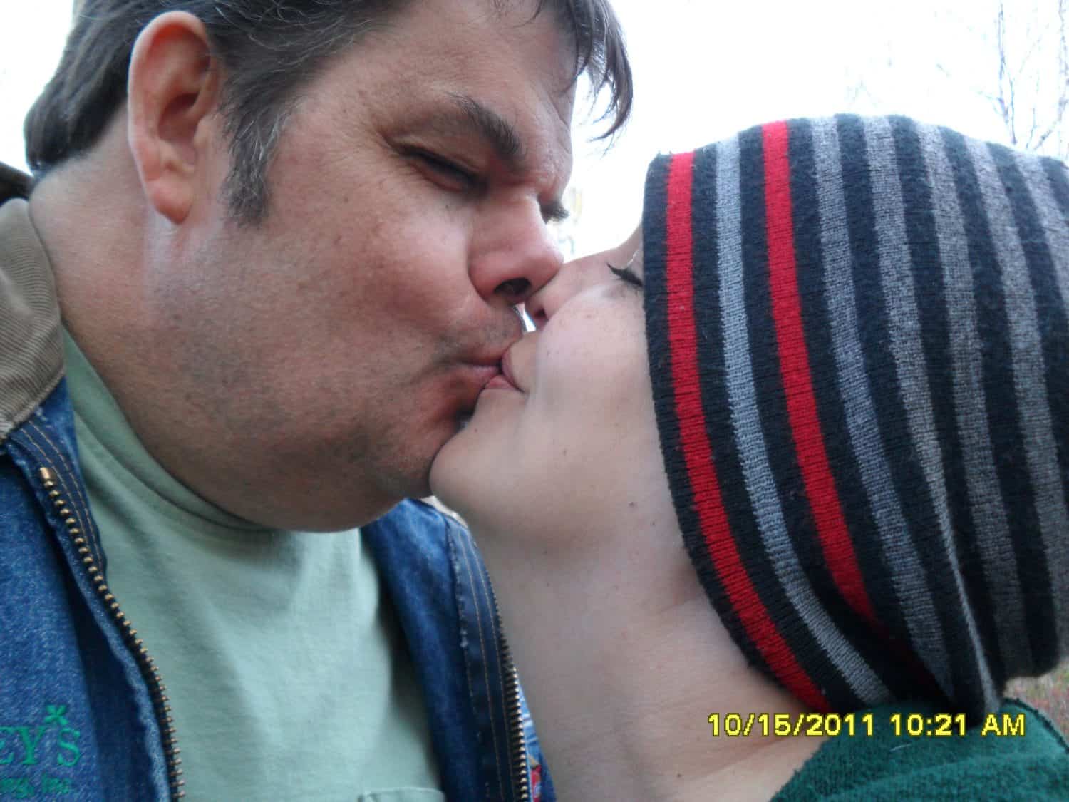 A close-up of two people sharing a kiss outdoors. One person wears a striped beanie while the other has short hair. The timestamp reads 10/15/2011 10:21 AM..